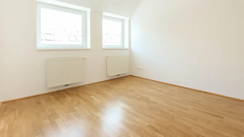 Helles Zimmer im Dachgeschoss mit Holzboden, zwei Fenstern und Heizkörpern.