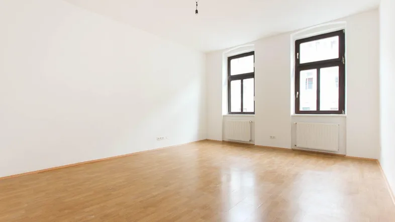 Helles, leeres Zimmer mit Parkettboden und zwei Fenstern, bereit zur individuellen Gestaltung.