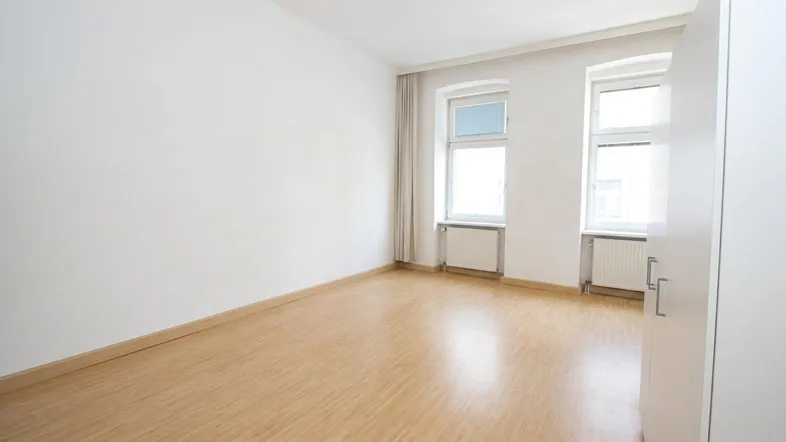 Helles, leeres Zimmer mit Laminatboden und zwei Fenstern, bereit zur individuellen Gestaltung.