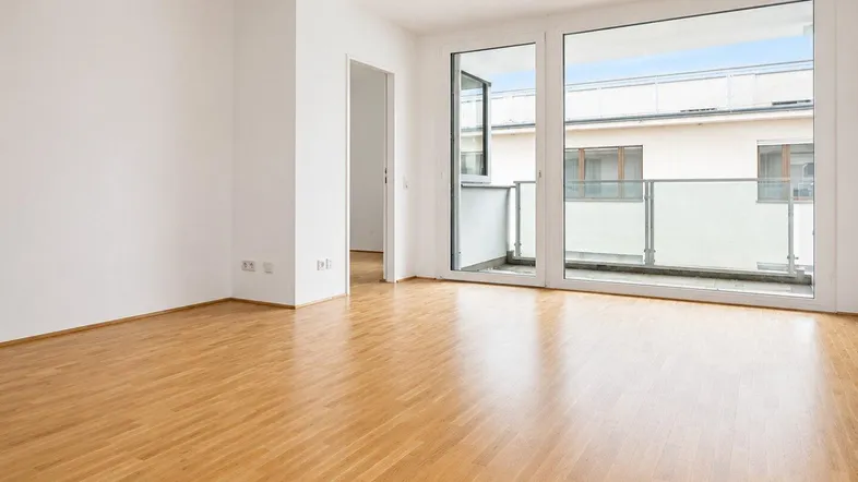 Helles Wohnzimmer mit Parkettboden, weißen Wänden und Zugang zu einem Balkon.