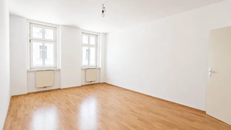 Helles, leeres Zimmer mit Laminatboden und zwei Fenstern, bereit zur individuellen Gestaltung.