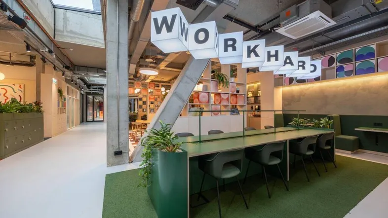 Moderner Coworking-Bereich mit langer grüner Arbeitsfläche, bequemen Stühlen und inspirierenden Leuchtschriftzügen.