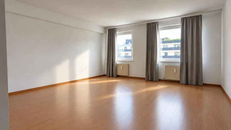 Helles, leeres Wohnzimmer mit Laminatboden und zwei Fenstern, die Tageslicht hereinlassen.