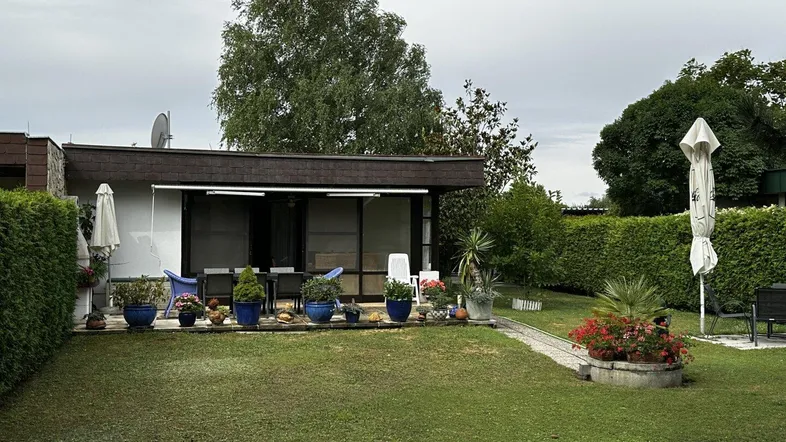 Großer Garten mit Rasenfläche und einer möblierten Terrasse vor dem Haus unter bewölktem Himmel.
