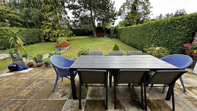 Große gepflasterte Terrasse mit Esstisch und Stühlen, umgeben von einem gepflegten Garten mit Hecken.