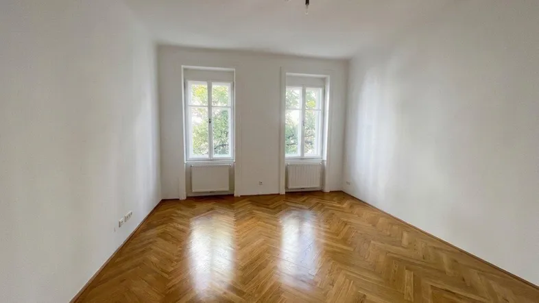 Helles Zimmer mit klassischem Fischgrätparkett und zwei Fenstern, ideal als Wohn- oder Schlafzimmer.