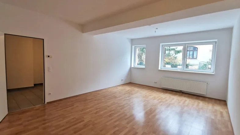 Geräumiges Wohnzimmer mit hellem Parkettboden und großen Fenstern, die viel Tageslicht hereinlassen.