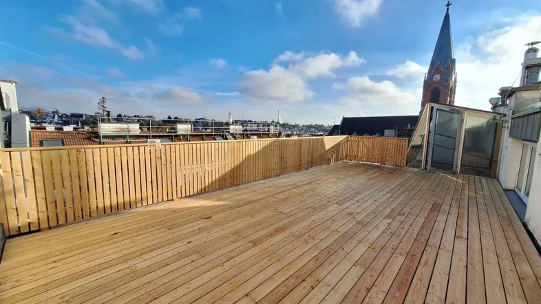 Weitläufige Dachterrasse mit frischem Holzdeck, Holzzaun und Blick auf die Stadt mit Kirchturm.