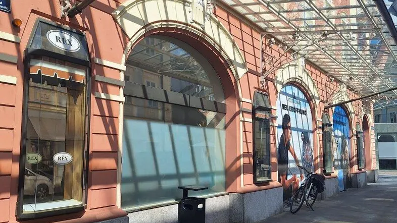 Historische Gebäudefassade mit großen Bogenfenstern und einem eleganten Glasvordach, beleuchtet von der Sonne.