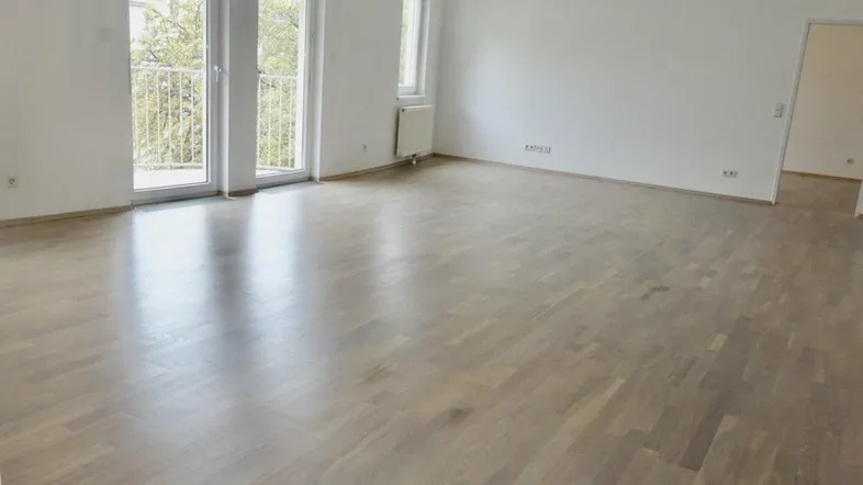 Helles, leeres Wohnzimmer mit Parkettboden und Zugang zu einem Balkon.
