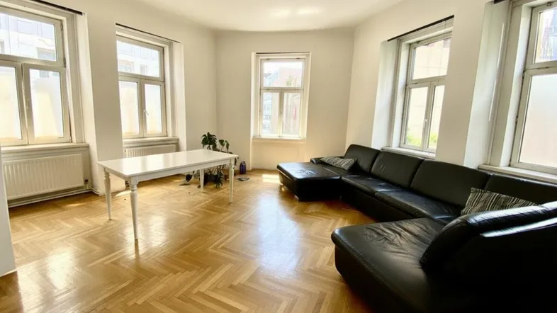 Helles Wohnzimmer mit Fischgrätparkett, großer schwarzer Couch und mehreren Fenstern.
