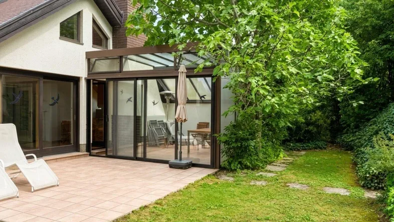 Gepflasterte Terrasse mit Liegestühlen und angrenzendem Wintergarten, umgeben von einem grünen Garten.