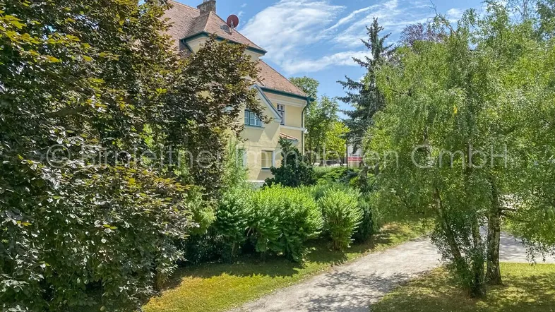 Historisches Gebäude mit klassischer Architektur, umgeben von altem Baumbestand und einem gepflegten Garten.