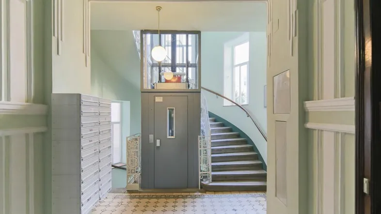 Helle Eingangshalle mit historischem Fliesenboden, modernem Aufzug und geschwungener Treppe.