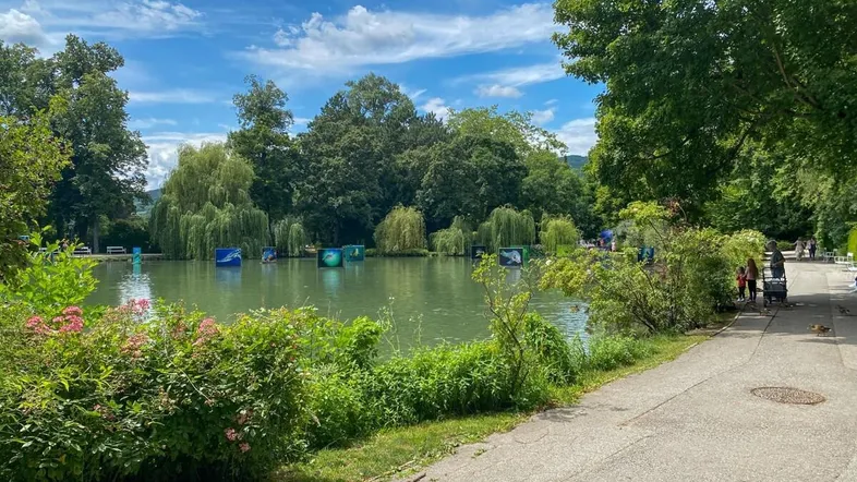 Seeufer mit Bäumen, einem Spazierweg und Kunstinstallationen auf dem Wasser unter blauem Himmel.
