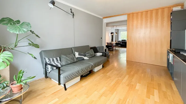 Helles Wohnzimmer mit Parkettboden, grauem Sofa und Pflanzen, offen zu einem weiteren Bereich.