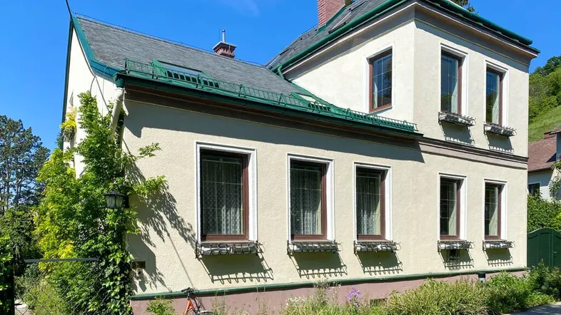Klassische Hausfassade mit mehreren Fenstern und gepflegtem Vorgarten unter blauem Himmel.