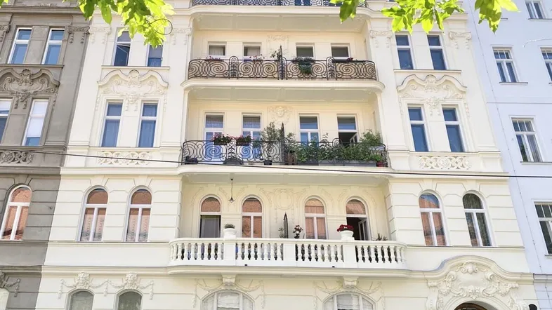 Elegante Altbaufassade mit reich verzierten Balkonen und großen Fenstern, umgeben von grünen Blättern.