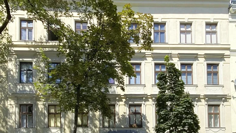 Historische Fassade eines Mehrfamilienhauses mit vielen Fenstern und Bäumen davor.