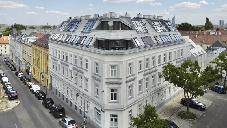 Gebäudeansicht eines Eckhauses mit modernem Dachausbau und vielen Fenstern in städtischer Umgebung.