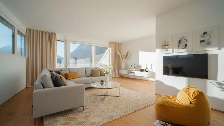 Helles und modern eingerichtetes Wohnzimmer mit bequemer Sitzecke, Fernseher und großen Fenstern mit Bergblick.