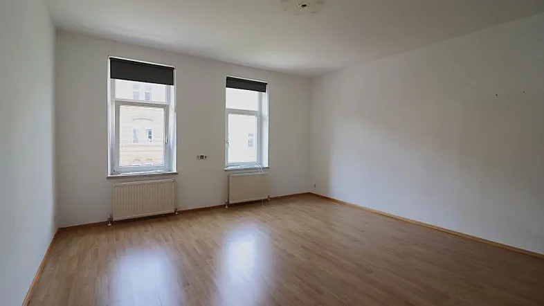 Helles, leeres Zimmer mit Holzfußboden und zwei Fenstern, bereit zur individuellen Gestaltung.