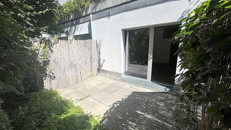 Eine gepflasterte Terrasse im Freien, umgeben von Grünpflanzen und einem Sichtschutz.