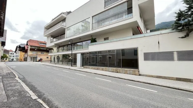 Modernes Gebäude mit großen Glasfassaden und Balkonen an einer Straße, umgeben von Bergen.