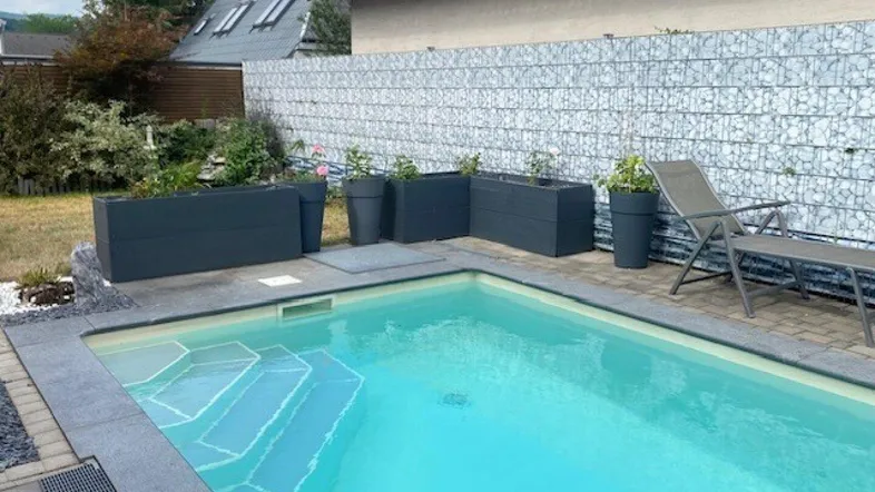 Moderner Außenpool mit Gabionenwand, umgeben von Pflanzen und einer Liegefläche.