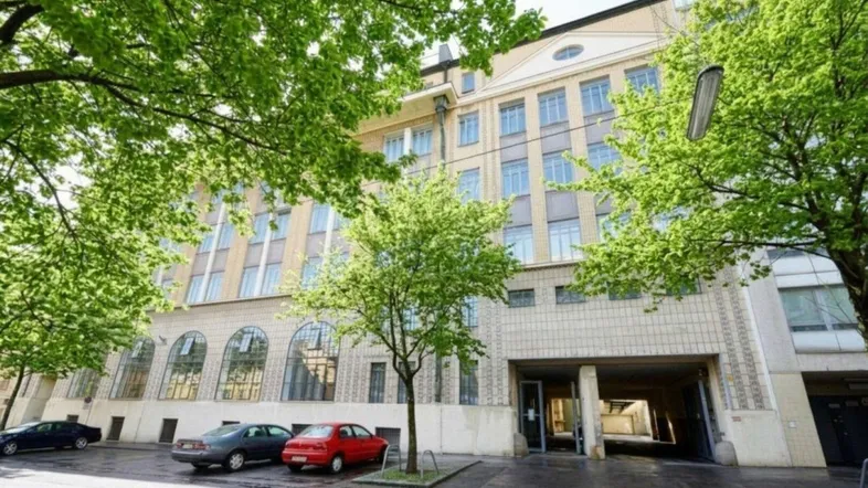 Gebäudefassade mit vielen Fenstern, umgeben von Bäumen und parkenden Autos auf der Straße.