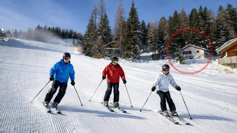 Skifahrer auf einer präparierten Piste mit einem Haus im Hintergrund, umgeben von verschneiten Bäumen.