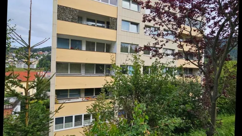 Mehrfamilienhaus mit gelber Fassade und Balkonen, umgeben von viel Grün und Hügeln.