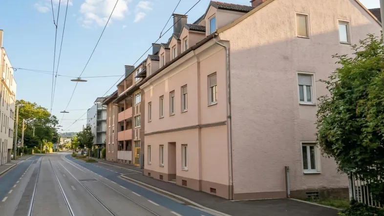 Ansicht der rosa gestrichenen Fassade eines Mehrfamilienhauses an einer städtischen Straße mit Straßenbahnschienen.