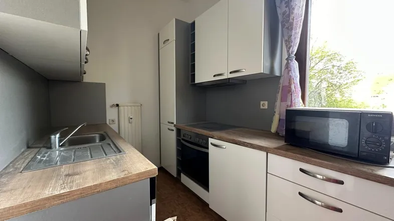 Kleine Küche mit weißen Schränken, Holzarbeitsplatte, Ofen und Mikrowelle am Fenster.