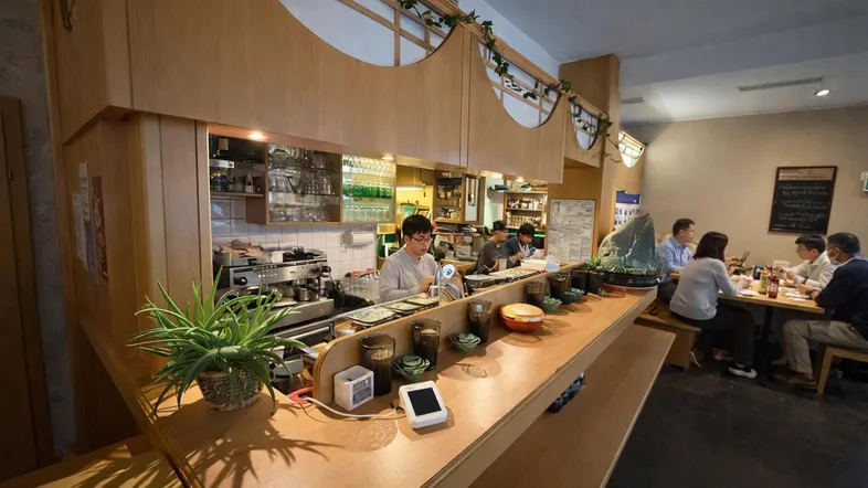 Innenansicht eines Restaurants mit Holzelementen, einer Theke und Gästen, die an Tischen sitzen.