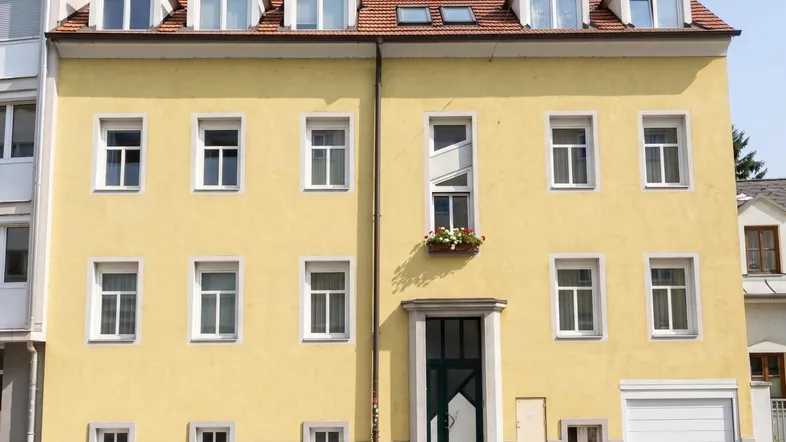 Gelbes Mehrfamilienhaus mit rotem Ziegeldach und mehreren Dachgauben in städtischer Umgebung.