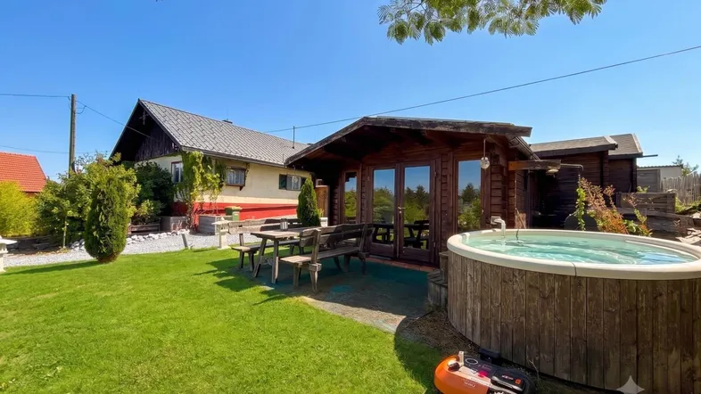 Gartenbereich mit Holzhaus, Whirlpool, Rasenfläche und Sitzgelegenheiten unter blauem Himmel.