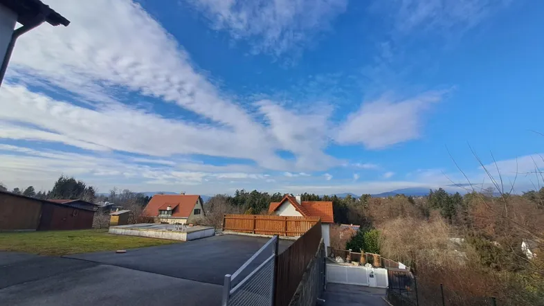 Weitläufiger Blick über eine Wohnsiedlung mit Häusern, Bäumen und Bergen im Hintergrund unter blauem Himmel.