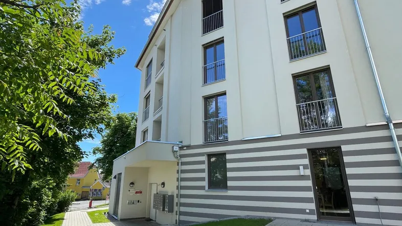 Moderne Wohnhausfassade mit kleinen Balkonen, gepflegter Grünanlage und hellem Eingangsbereich.