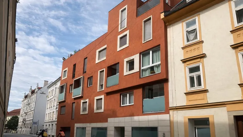 Modernes Gebäude mit markanter roter Fassade und unregelmäßig angeordneten Fenstern in urbaner Umgebung.