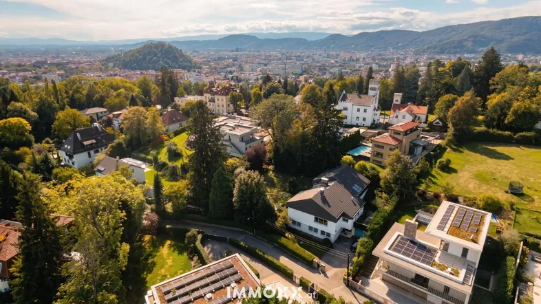 Panoramablick auf eine grüne Wohngegend mit vielen Häusern und Bergen im Hintergrund.