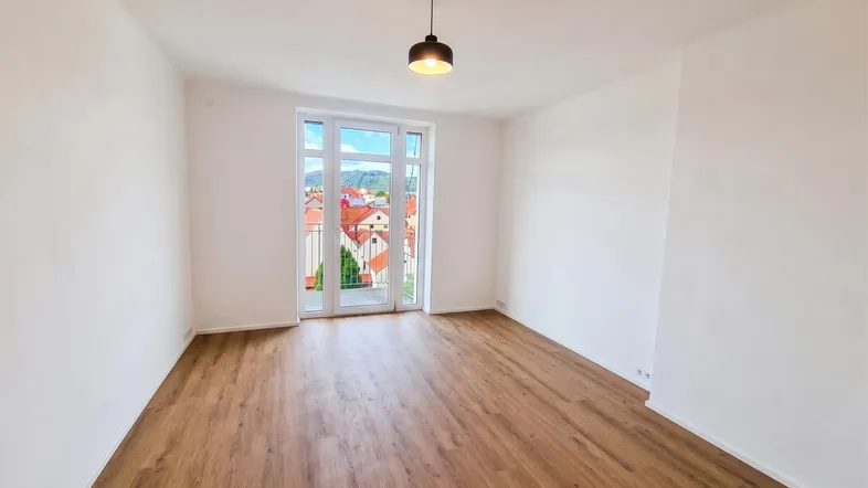 Helles Zimmer mit Holzboden und Zugang zu einem kleinen Balkon mit Blick auf die Stadt und Hügel.