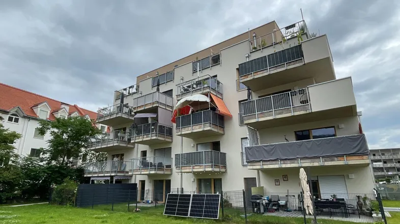 Modernes Mehrfamilienhaus mit mehreren Balkonen und einer Grünfläche im Vordergrund unter bewölktem Himmel.