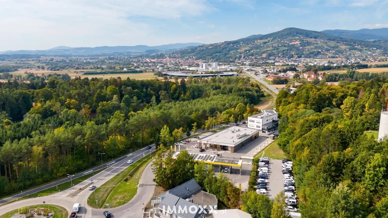 Luftaufnahme eines Gewerbegebiets mit Straßen, viel Grün und Bergen im Hintergrund unter blauem Himmel.
