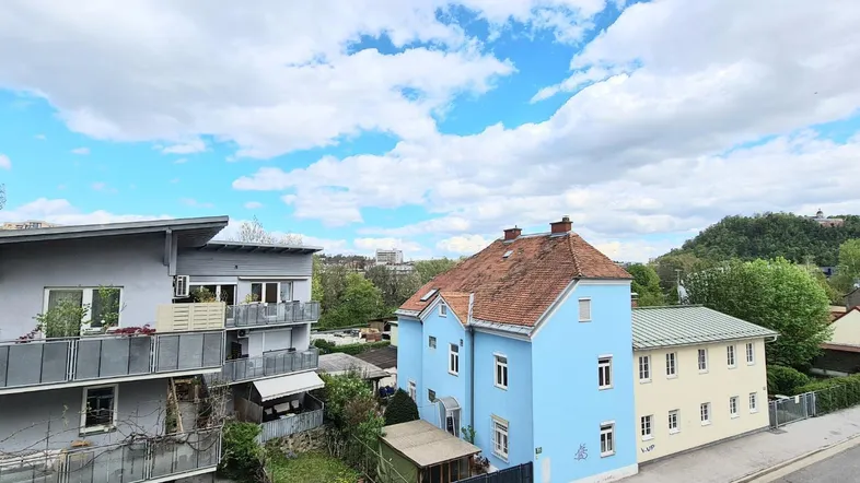 Blick auf mehrere Gebäude mit blauer Fassade und rotem Dach sowie eine grüne Hügellandschaft im Hintergrund.
