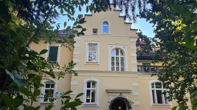 Historisches Gebäude mit gelber Fassade und verspielten architektonischen Details, umgeben von üppigem Grün.