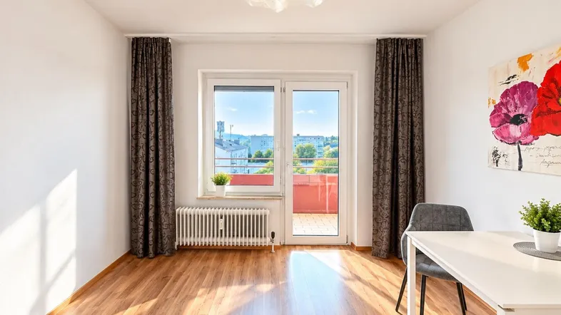 Helles Zimmer mit Holzboden, Zugang zum Balkon und einem kleinen Essbereich.