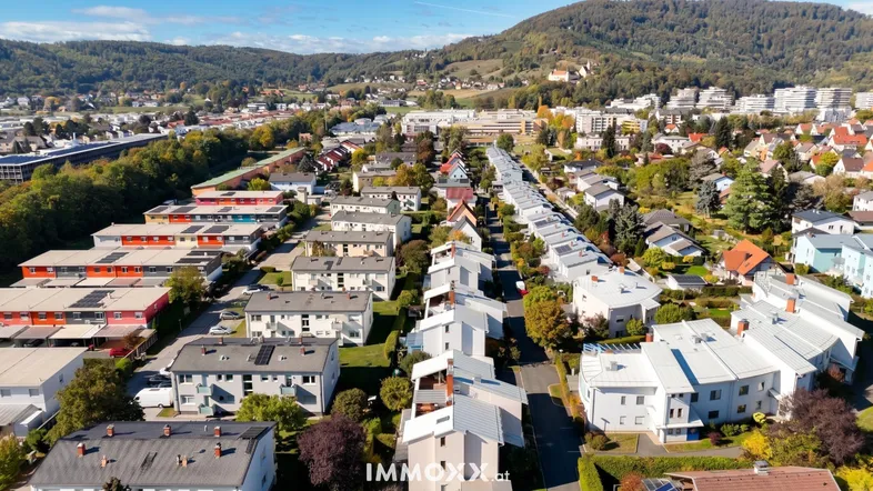 Luftaufnahme einer Wohnsiedlung mit vielen Häusern, grünen Gärten und bewaldeten Hügeln im Hintergrund.