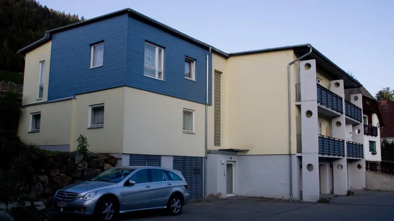 Gebäudeansicht mit blauer Holzverkleidung und gelber Fassade, ein Auto parkt davor.