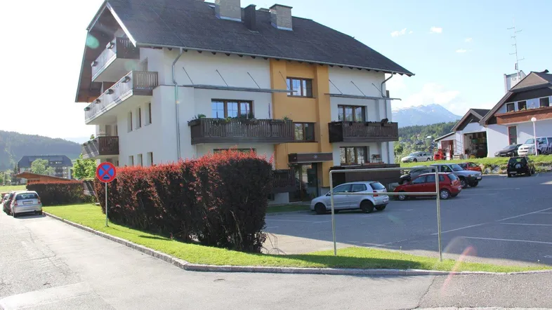 Mehrfamilienhaus mit traditioneller Architektur, Balkonen und Parkplätzen im Freien, umgeben von grüner Landschaft und Bergen.